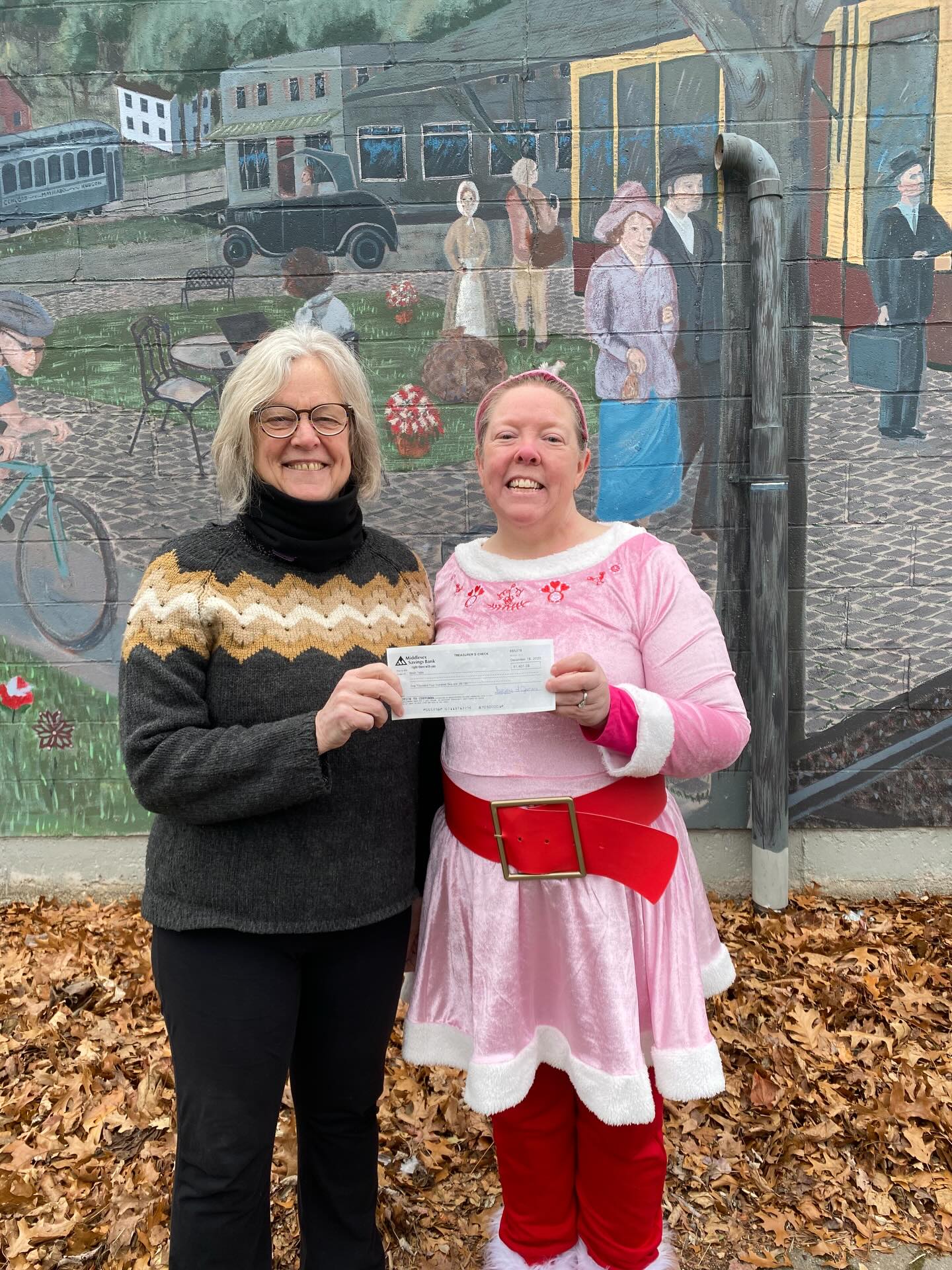 Two women holding a check between them. One of them is in a pink elf costume, the other in a black sweater and pants.