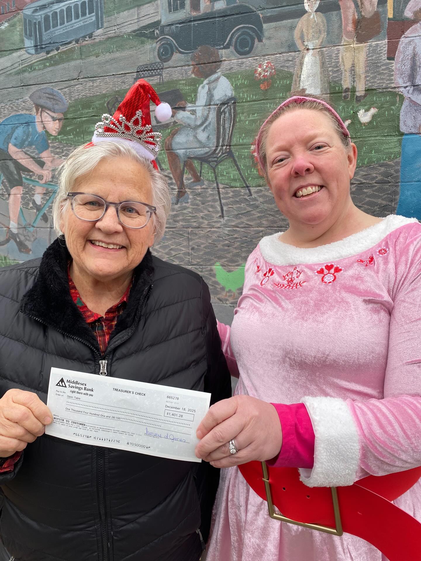 Two woman holding a check between them, one in a pink elf costume, the other in a black jacket with santa hat