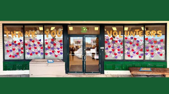 Picture of the front windows at Open Table with colored paper hearts that have volunteer names on them and painted words Thank You Volunteers above
