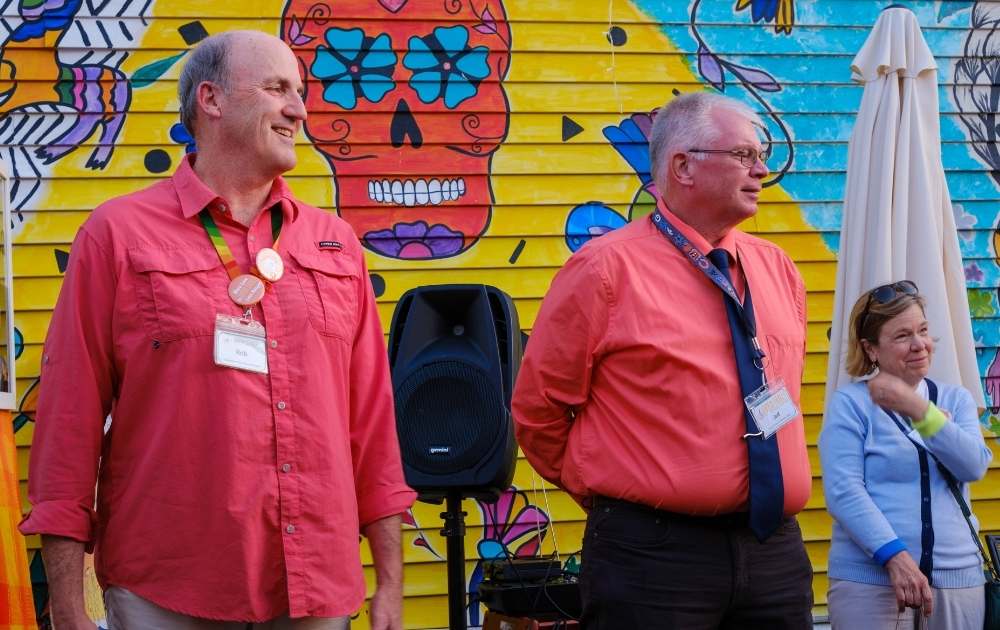 Two white males in orange shirts standing in front of mexican sugar skull mural